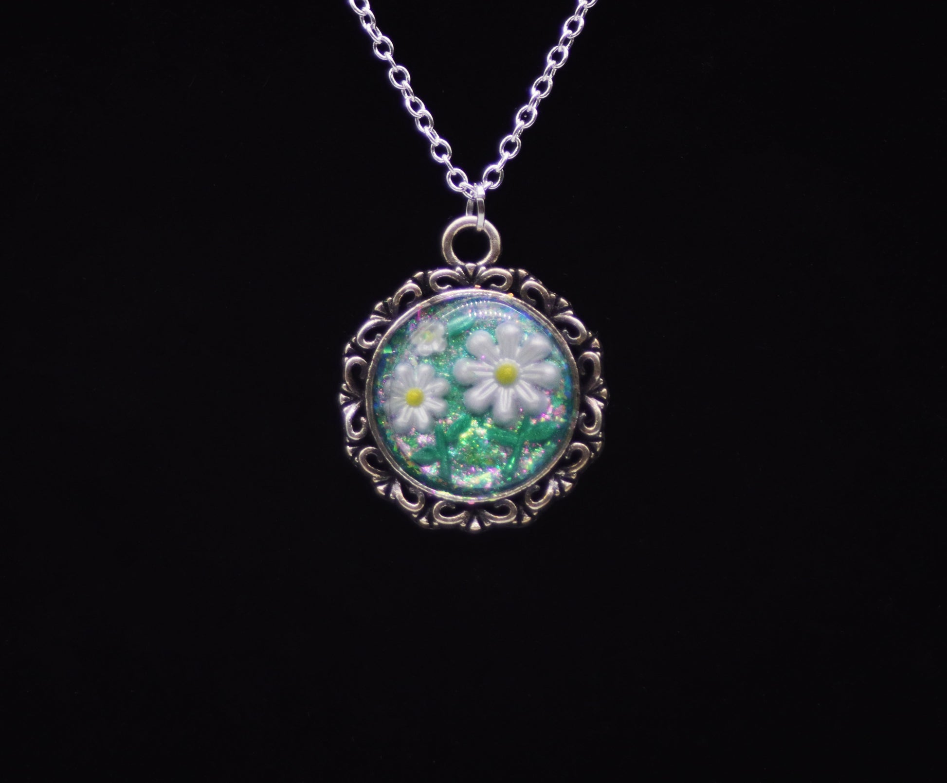 White resin daisy necklace with green glitter background in silver-tone round filigree frame on silver chain against black background